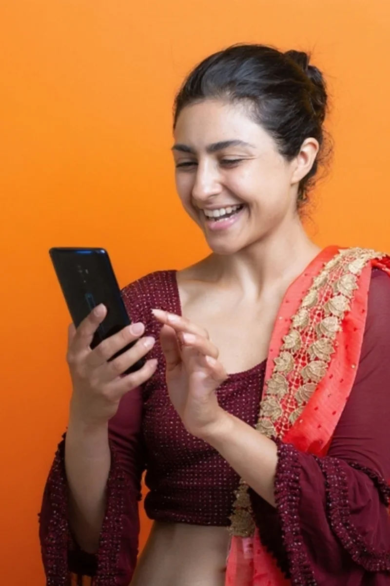 A woman in a maroon kurta smiles at her phone, repeating a phrase aloud