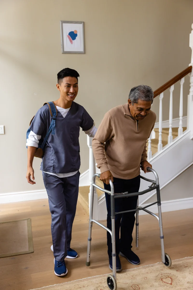 A carer in scrubs walks beside an elderly man using a walking frame at home