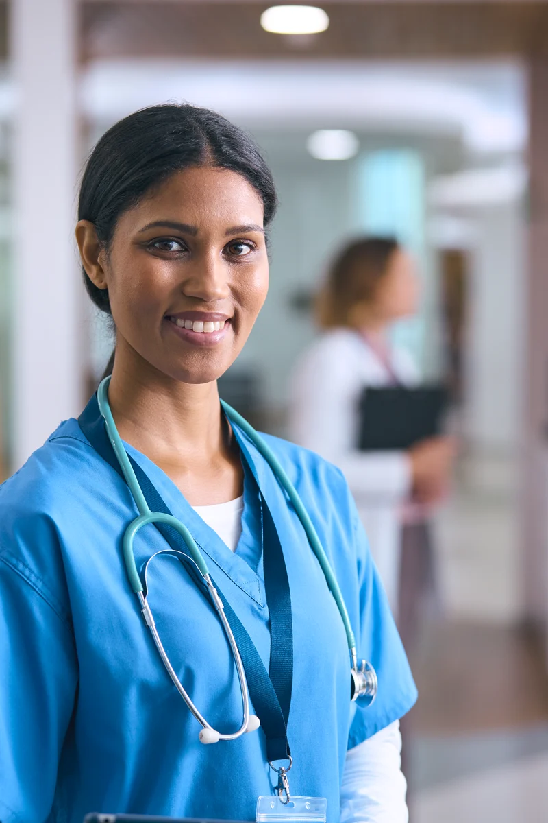 A nurse in scrubs with a stethoscope stands in a hospital corridor