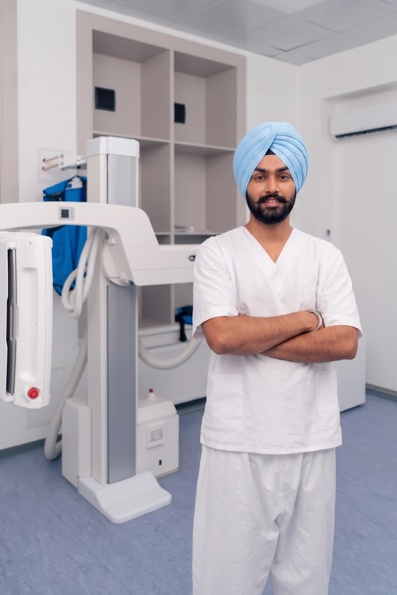 A radiographer in white scrubs stands beside an X-ray unit in an imaging room