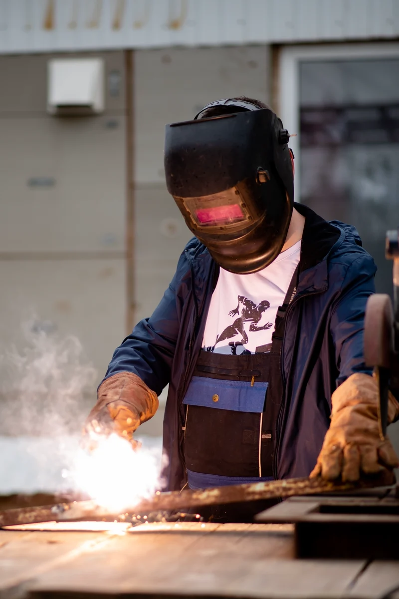 A welder in a full face shield lays a bead on a steel plate, sparks falling, in a workshop